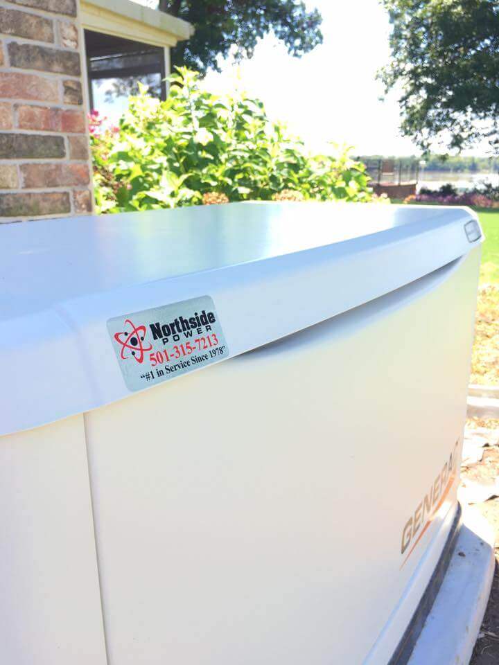 A white whole-home generator sits outdoors by a brick wall, partially in shade. A sticker reads Northside Power, with a phone number and notes In Service Since 1978. Greenery surrounds the scene, an ideal setup for disaster preparedness as tornado season approaches.