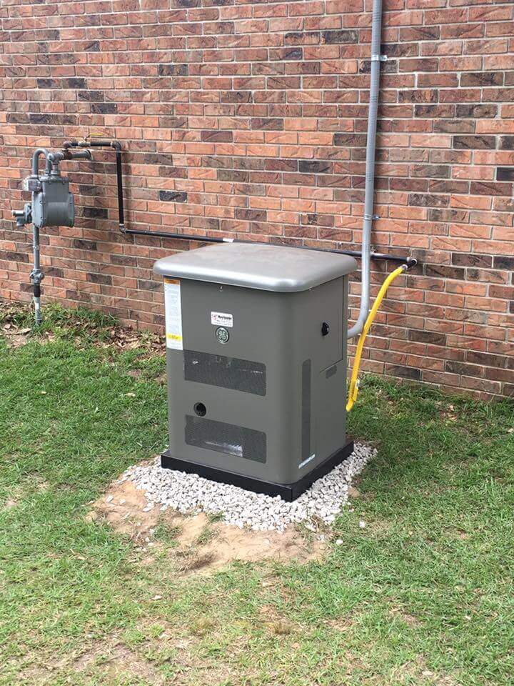 A gray whole-home generator with a yellow hose is installed on a concrete pad surrounded by small white rocks. Positioned next to a brick wall with visible piping and a gas meter, its an essential part of disaster preparedness during tornado season, amidst the grassy surroundings.