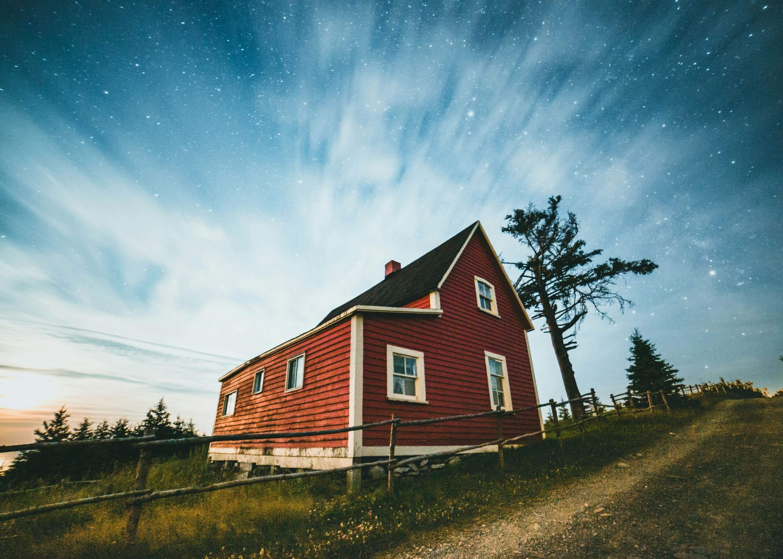 A red wooden house sits beside a dirt road under a starry night sky with wispy clouds, surrounded by trees and grass.