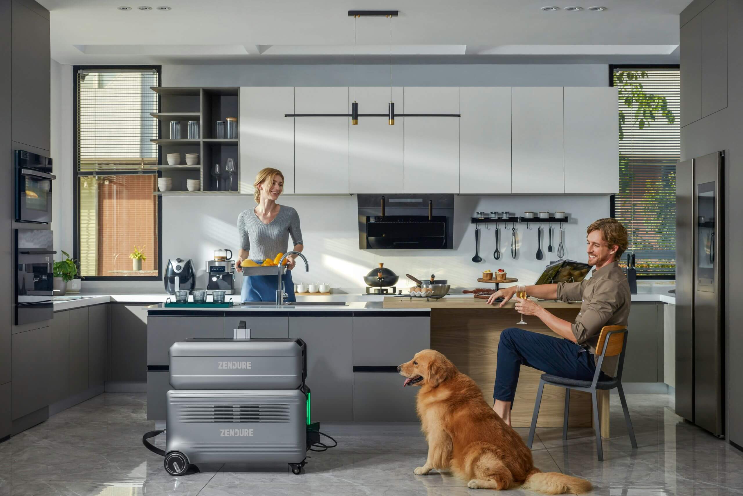 A woman cooks in a modern kitchen while a man sits nearby, petting a golden retriever. Portable power stations are on the floor. The room is bright with natural light and minimalist decor.