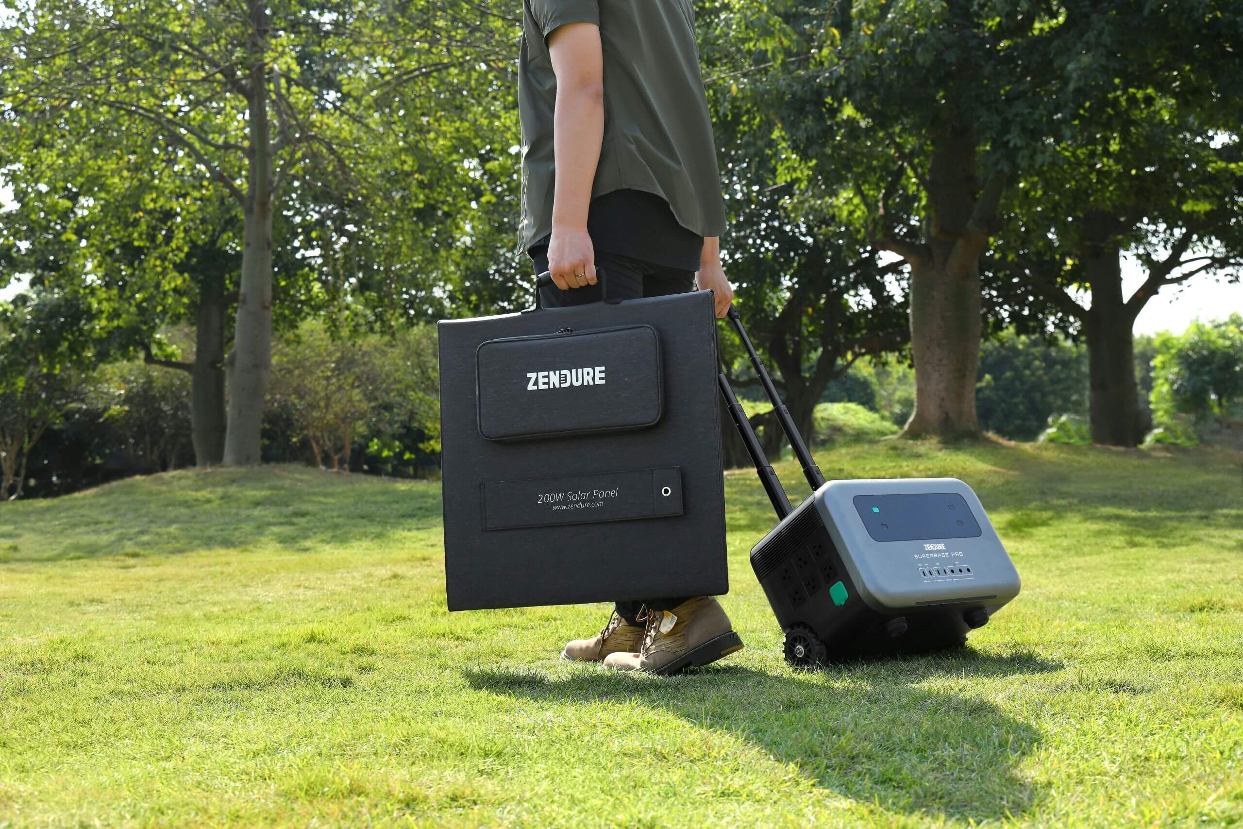 A person carries a folded Zendure solar panel while pulling a rolling Zendure portable power station across a grassy park.