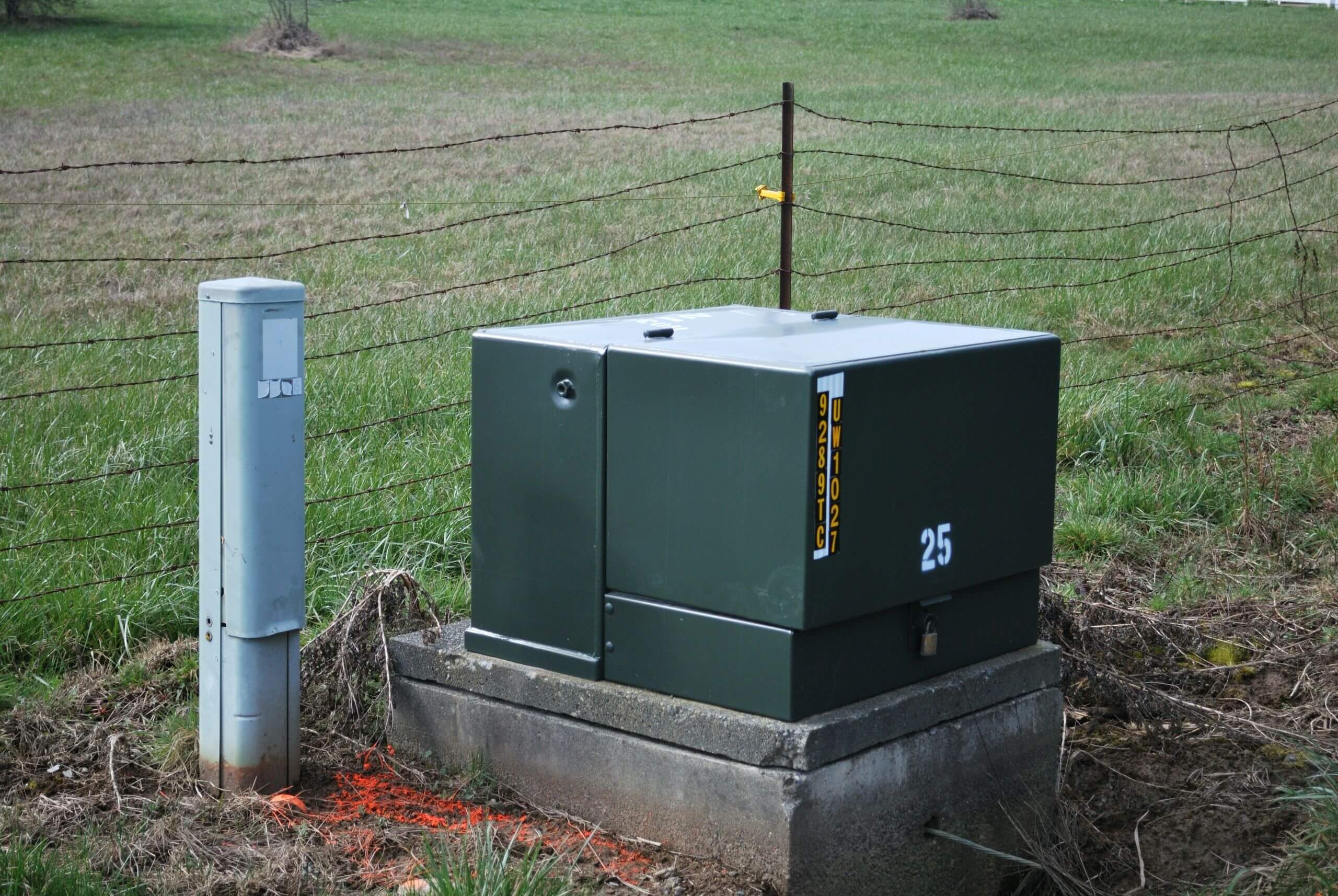 A dark green electrical transformer box labeled with the number 25 and serial number UW1027, standing on a concrete pad next to a utility post in a grassy field.